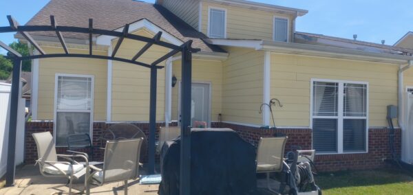 Backyard patio under a dark metal pergola with beige outdoor chairs and a covered grill in front of a yellow house with brick trim.