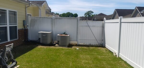 Backyard with two outdoor AC units along a white privacy fence, yellow house, and a hose reel in front left.