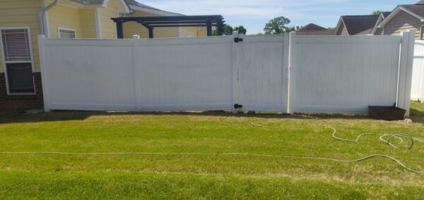 Backyard with a white vinyl privacy fence and a closed gate, yellow house siding visible on the left, and a coiled hose resting on the green lawn.