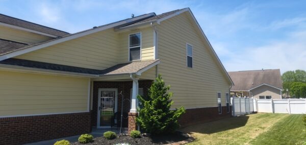 Two-story yellow house with brick foundation, white trim, and a small evergreen near the front porch on a sunny day.