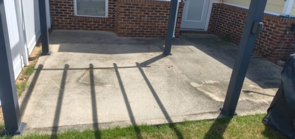 Concrete patio under a dark metal pergola, brick house in background and long vertical shadows across the concrete surface in sunlight.