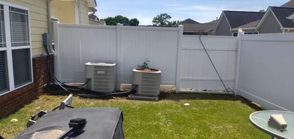 Backyard with two outdoor air conditioning units along a white fence, beside a yellow house with brick foundation.