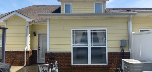 Rear view of a yellow house with brick foundation, a large white-framed window with blinds, and a side door.