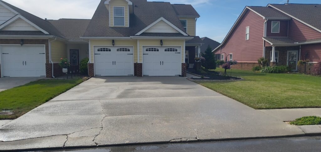 Suburban street scene with a yellow house in the center, three white garage doors, brick accents, and manicured lawns on either side.