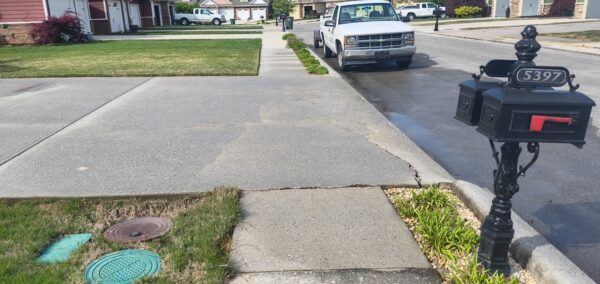 Suburban street scene with a white pickup parked along the curb, a decorative black mailbox labeled 5397, green lawns, and a cracked concrete sidewalk; a small planting bed and storm drain covers are nearby.