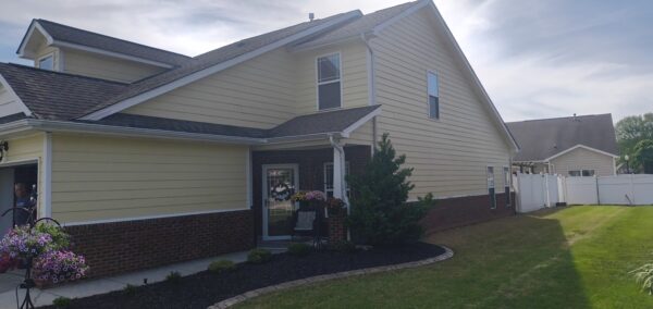 Front view of a beige two-story house with brick foundation, small porch, and potted flowers by the entrance with a white privacy fence in the background.