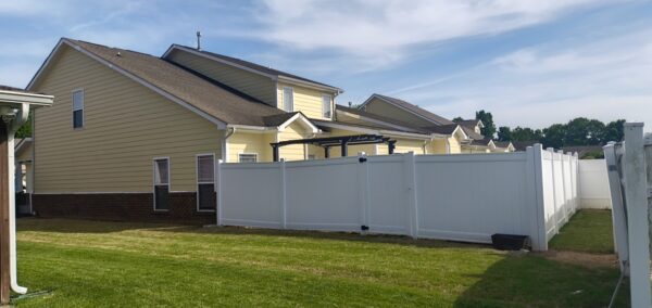 Backyard scene with yellow house, brick foundation, white privacy fence, and a black pergola under a blue sky.