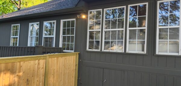 Gray wooden house exterior with white-trimmed, multi-pane windows and a wooden privacy fence in the foreground.