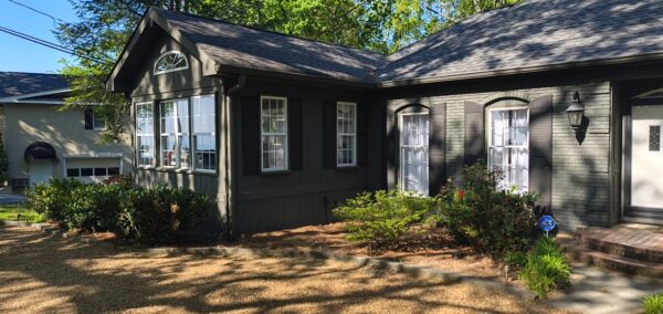 Dark gray, cottage-style house with multiple white-framed windows and shutters, front garden in bloom