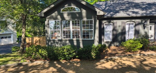Gray house with large bay window and arched transom, black shutters, and a garden bed of green shrubs along a gravel path.