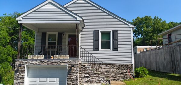 Two-story house with a stone lower facade, white siding, black shutters, a red front door, and a small dog on the front steps.