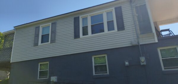 Side view of a two-story house with gray siding, white trim, and dark shutters against a clear blue sky, with a deck on the left.