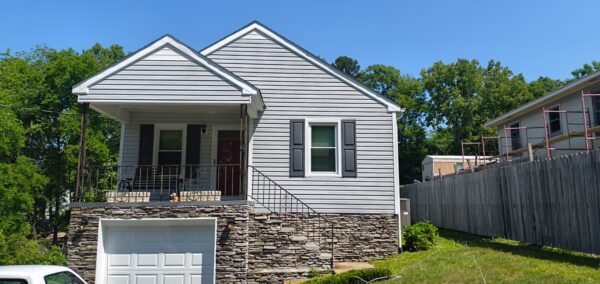 Two-story house with gray siding, stone foundation, front porch, and attached garage underneath the living area.