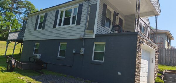 Two-story house with a gray lower level, light siding, dark shutters, and a stone-faced garage door; a flatbed trailer rests on the lawn to the left.