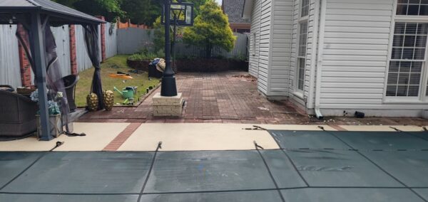Backyard patio with brick pathway, a pool cover in the foreground, and a white house with windows on the right side. A gazebo and various garden toys sit along a wooden fence line in the background.