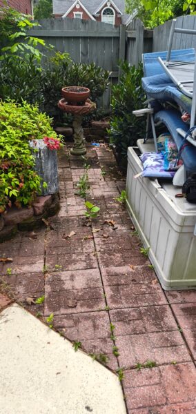 Narrow brick patio in a backyard with weeds growing between bricks, a pedestal planter near center, and stacked blue outdoor chairs to the right against a fence.