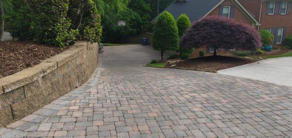 Curved brick-paver driveway with a stone retaining wall on the left and landscaped trees on the right, leading to a house in the distance.