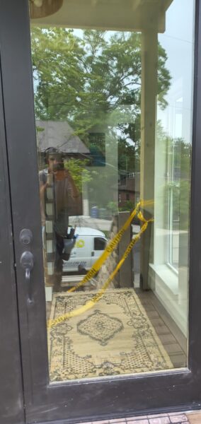 Glass front door with bright yellow caution tape across the entry; reflection of the photographer in the glass and a patterned rug inside.