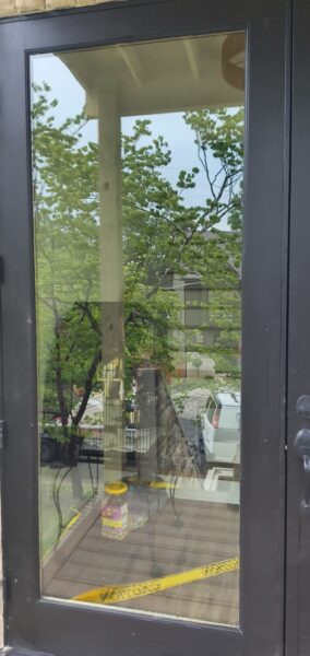 Front glass door with a black frame reflecting green trees outside; yellow caution tape spans the interior floor near a small container by the threshold.
