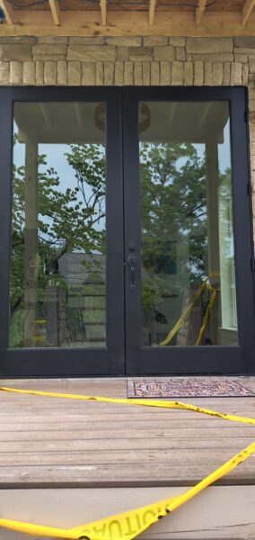 Double glass doors with black frames set in a stone facade, yellow caution tape across a wooden deck in front; interior shows construction materials and equipment.