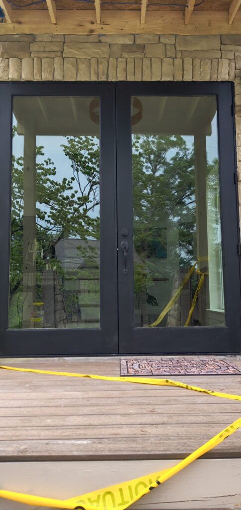 Double glass doors with black frames set in a stone facade, yellow caution tape across a wooden deck in front; interior shows construction materials and equipment.