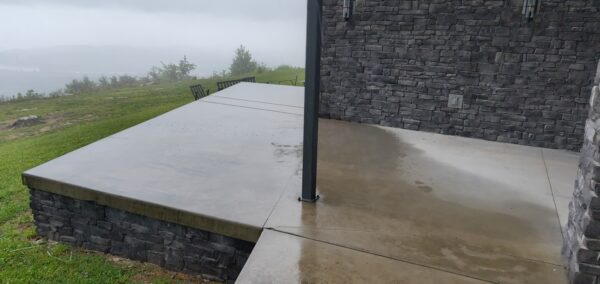 Wet concrete patio attached to a stone building on a misty hill, with a metal post and distant outdoor chairs.