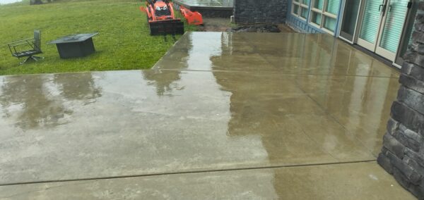 Wet concrete patio beside a house with glass doors, reflecting a rainy sky after rainfall in the surface. Object on the lawn include a black box-like table and an orange lawn tractor in the distance.