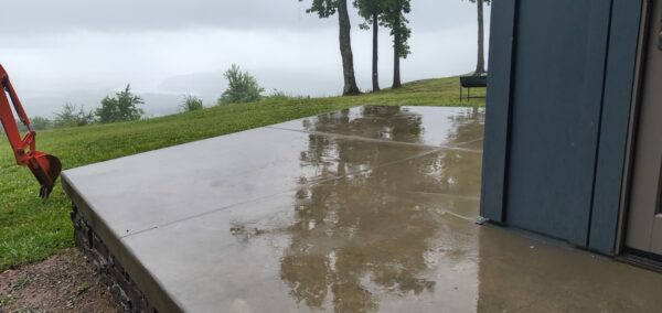 Wet concrete patio outside a blue building, reflecting trees on a misty hillside with an orange excavator arm visible on the left.