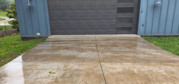 Wet concrete driveway leading to a dark gray garage door set in a blue metal building; green lawn visible on both sides.