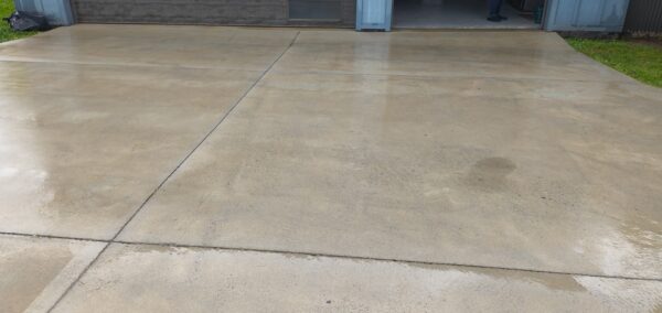 Wide concrete driveway leading to an open garage; wet patches on the surface and a person standing inside near the doorway.