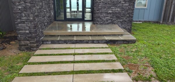 Front entry with dark stone walls, glass door, and a series of wide concrete steps leading to the door, flanked by grass.