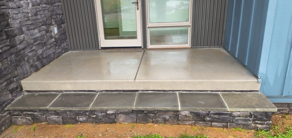 Concrete entry landing with two wide steps in front of a glass door, flanked by stone veneer walls and blue metal siding.