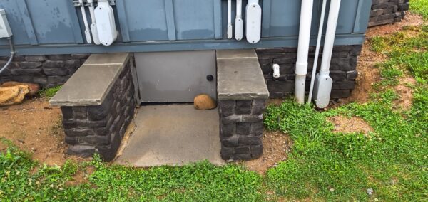 Outdoor utility access area with a metal door set into stone block pedestals and a rock on the step.