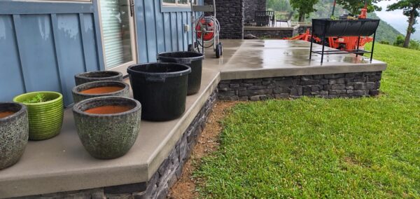 Row of ceramic and green pots filled with red soil on a concrete porch beside a blue house siding and stone foundation, with a grassy yard beyond.