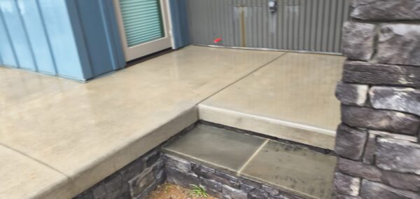 Beige concrete porch with glass door, blue siding to the left, and a stacked stone wall on the right; a small red object sits on the upper landing edge.