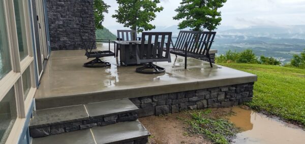 Wet concrete patio with four black chairs around a square table on a stone deck, overlooking a valley.