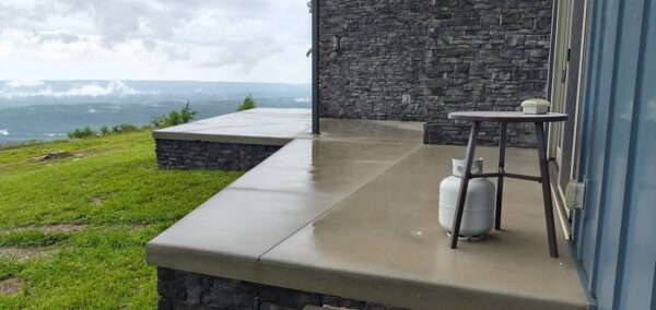 Rainy concrete patio beside a stone wall, propane tank and small round table on a hillside overlooking rolling green hills and a distant valley.