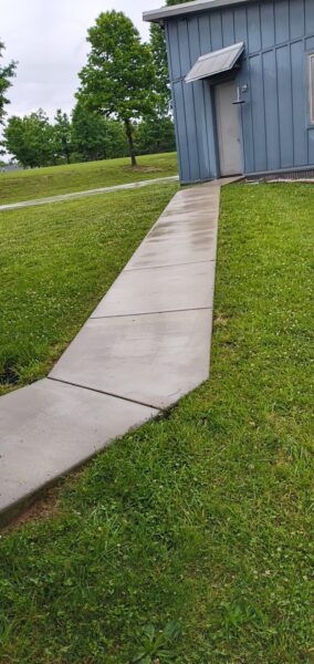 Concrete walkway leading to a blue metal building entrance with a small overhang, surrounded by green grass and trees in a park-like setting.