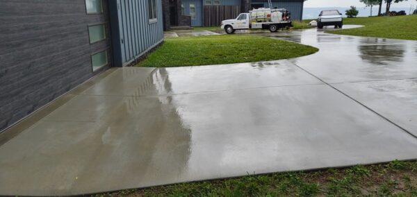 Wet concrete driveway in front of a modern house after rain, with a curved path and green lawn nearby.