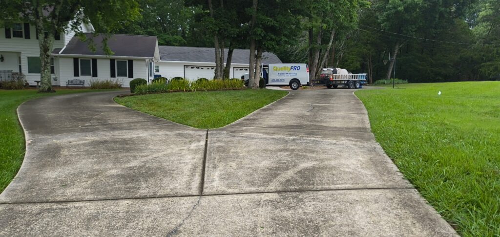 Split concrete driveway curves toward a white house with a 'QualityPRO' power-washing van and trailer near trees on a green lawn edge.