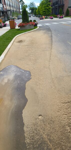 Sidewalk and street with planters and flowers; a sandy spill covers part of the pedestrian path from curb to the road.
