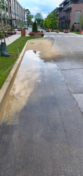 A street with wet pavement and a large puddle along the curb, reflecting the cloudy sky and nearby trees and buildings; planters line the sidewalk in a modern development.