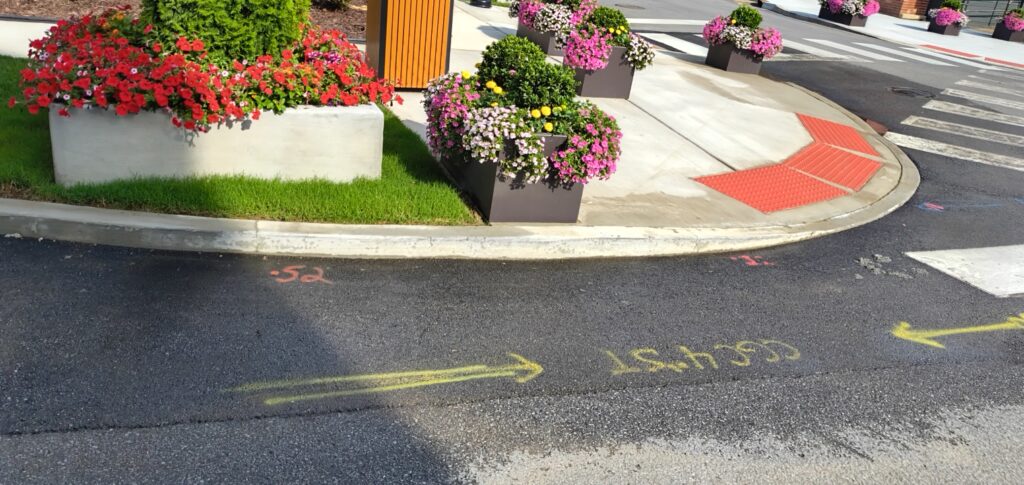 Decorative curb area with colorful flower planters and a sidewalk, showing yellow directional arrows painted on the road and red tactile paving near the curb edge used for pedestrian guidance.