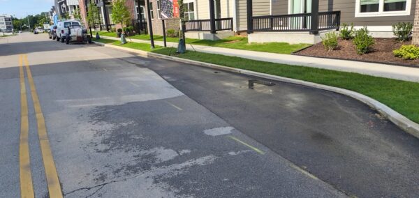 Residential street with fresh asphalt patch along the curb, parked vans, and a sidewalk border with new landscaping nearby